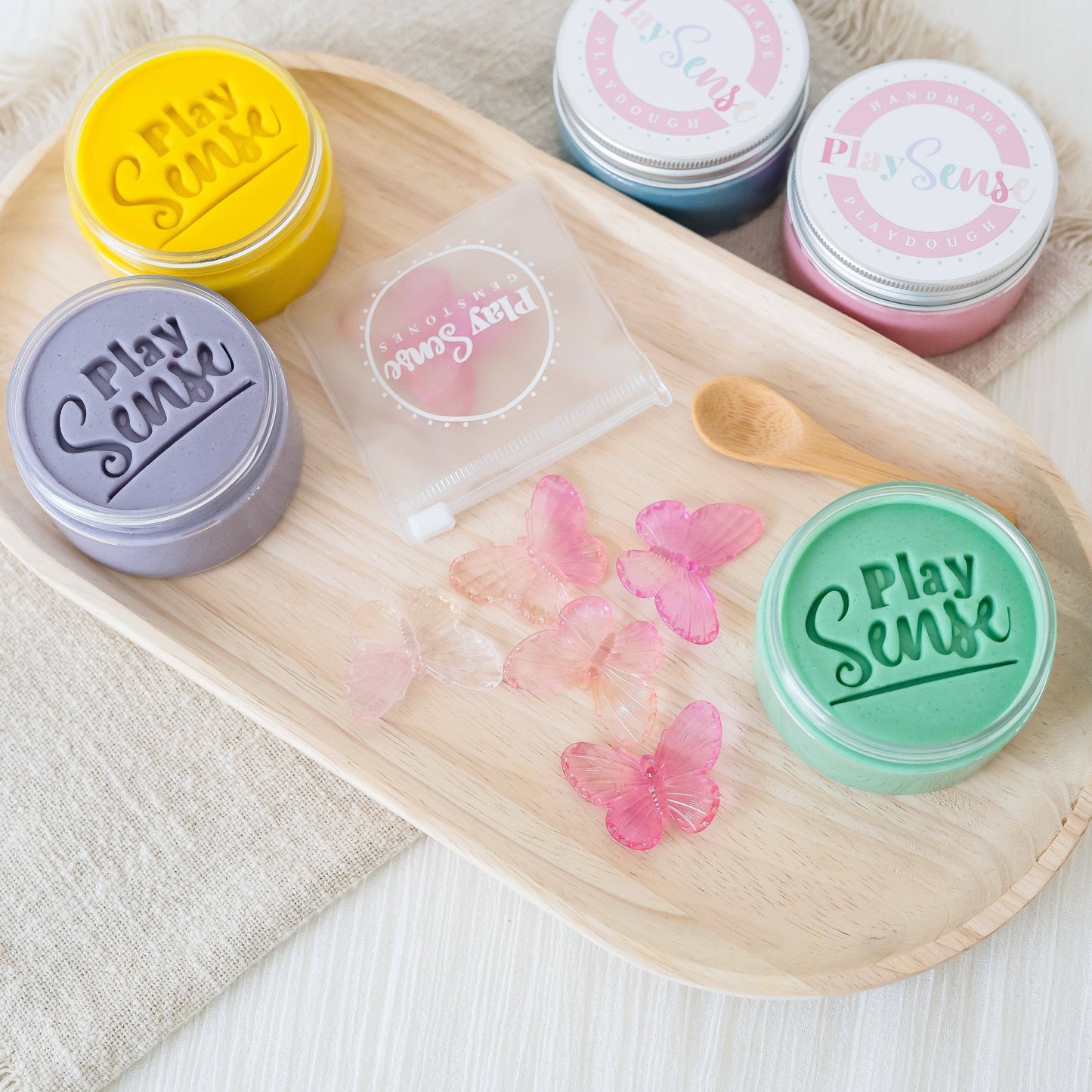 Multiple containers of Play Sense play dough with colorful gemstone butterflies displayed on a wooden tray.