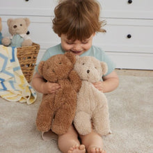 Child holding two teddy bears in a room with a basket of more teddy bears and a blanket.