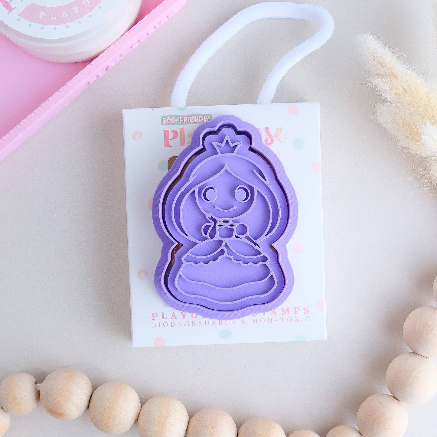 A playdough stamp with a princess shape design, displayed on a card with the Play Sense brand details. The stamp is purple, and there are wooden beads and a pink tray in the background.