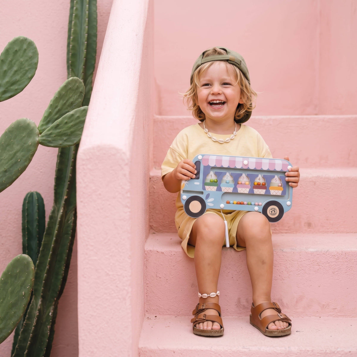 Child holding a wooden ice cream puzzle toy against a pink wall with a cactus plant.