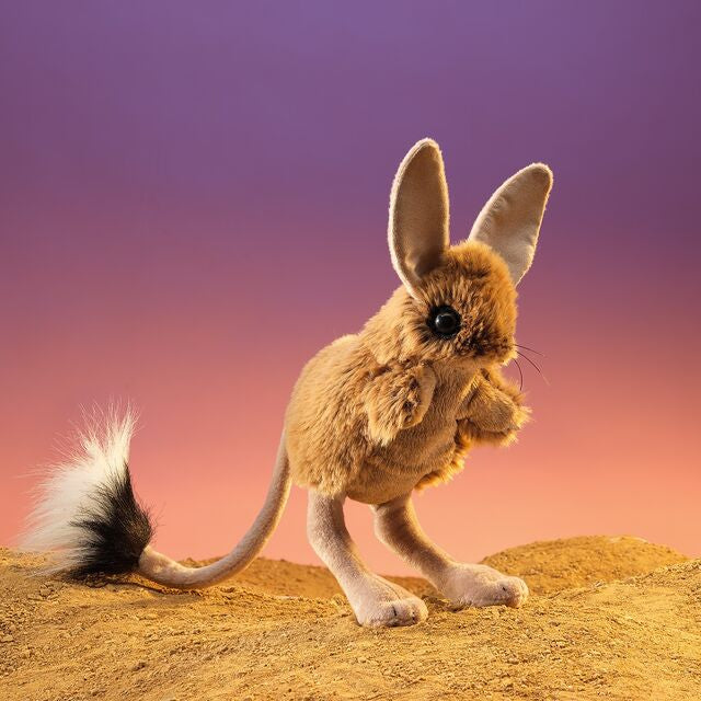 Plush toy of a long-eared jerboa on a sand dune with a colorful sky background