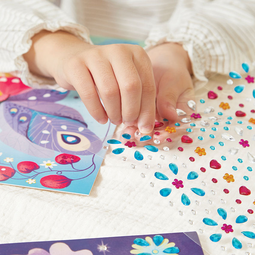 Child's hand decorating a craft project with colorful gem stickers on a white surface.