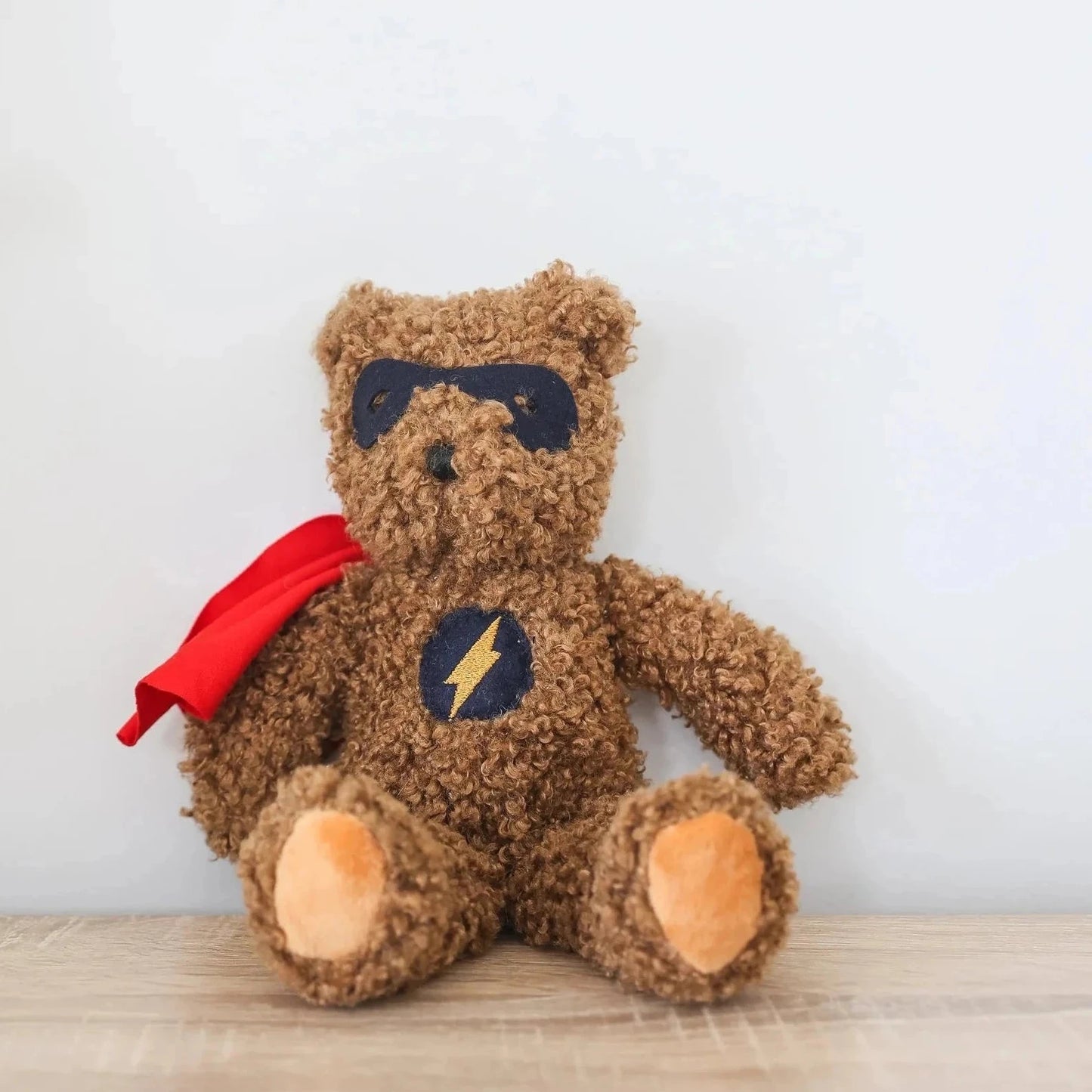 Teddy bear with a superhero cape on a wooden surface with a white wall background