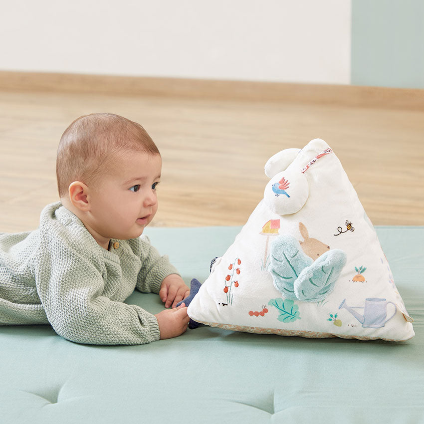 Baby lying on a bed next to a soft pyramid-shaped pillow with cartoon characters