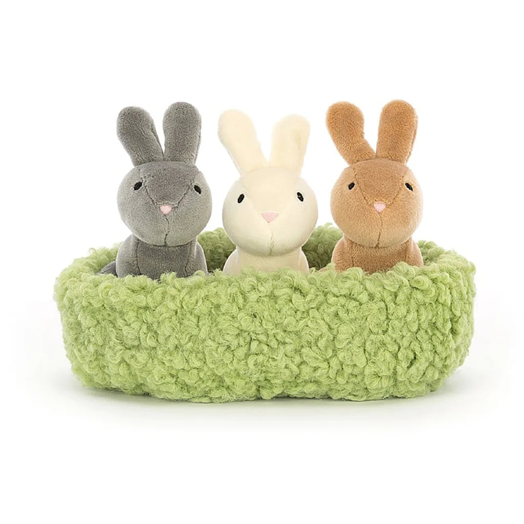 Three plush toy rabbits with perky ears, placed in a fluffy green nest. One is grey, one is white, and one is brown.