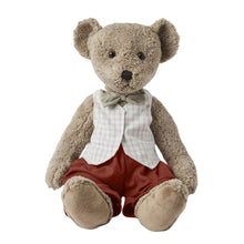 A large plush teddy bear wearing a white checkered vest and red pants with a green bow-tie, sitting upright.