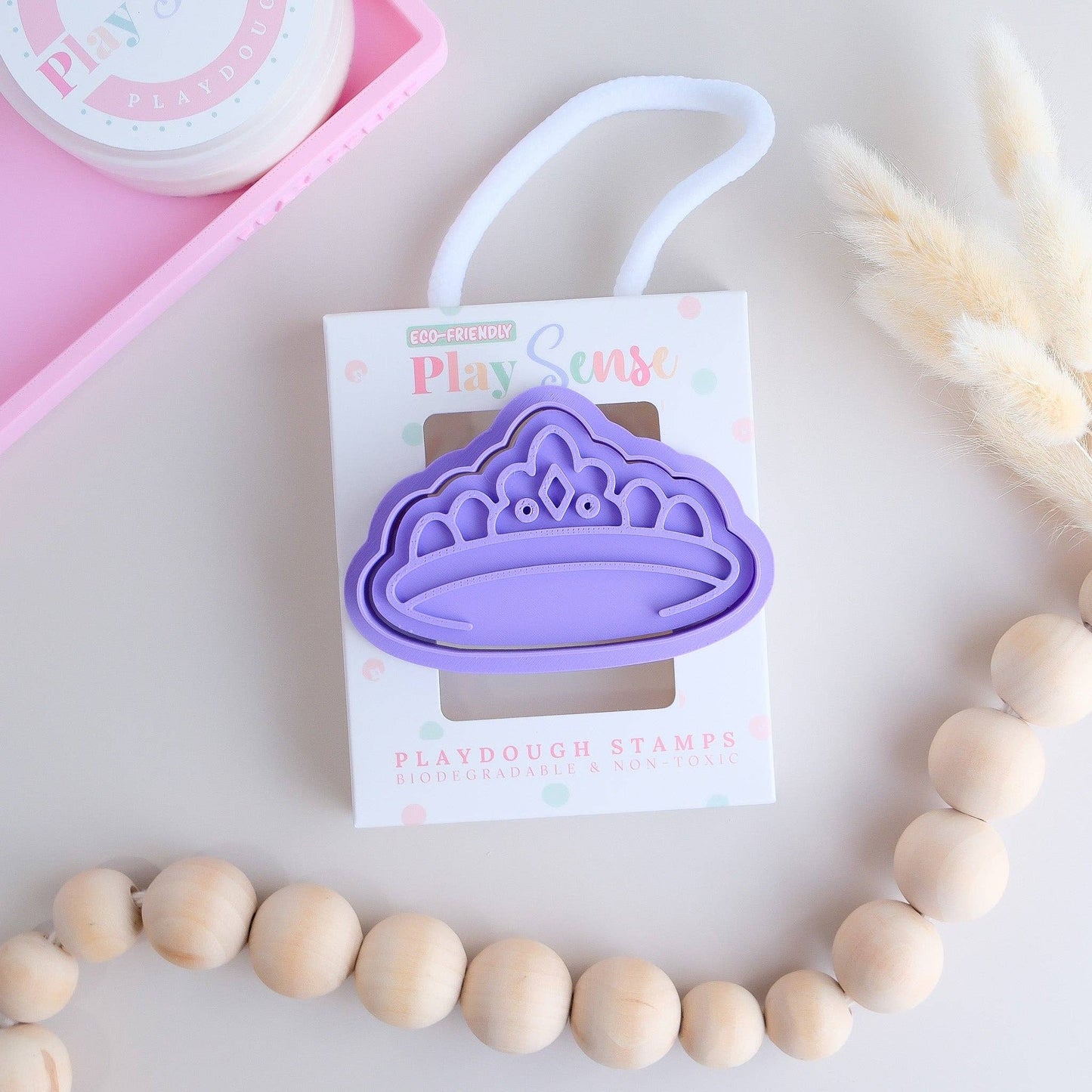 A purple crown-shaped playdough stamp on a packaging card, with a white handle and decorative elements in the background.
