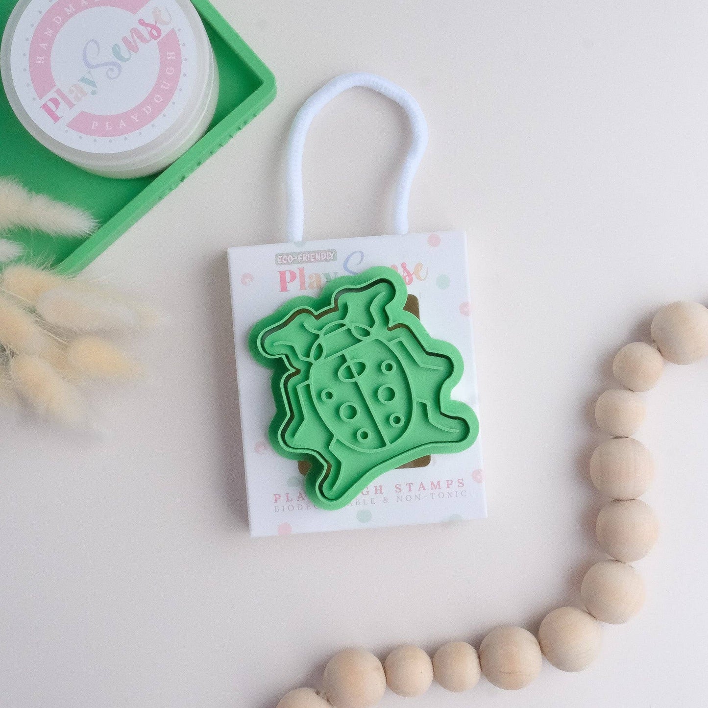 A green ladybug-shaped playdough stamp on a white card with the Play Sense brand name, accompanied by a container and decorative elements in the background.