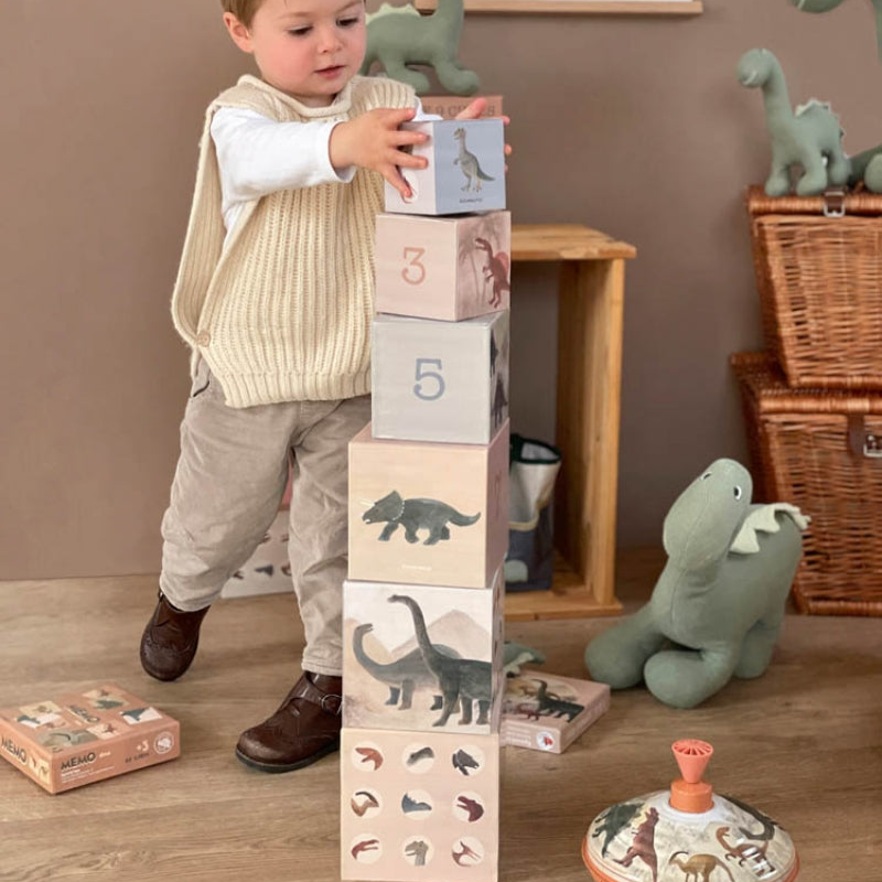 Child playing with cardboard stacking blocks featuring dinosaur illustrations in a room with toy dinosaurs.