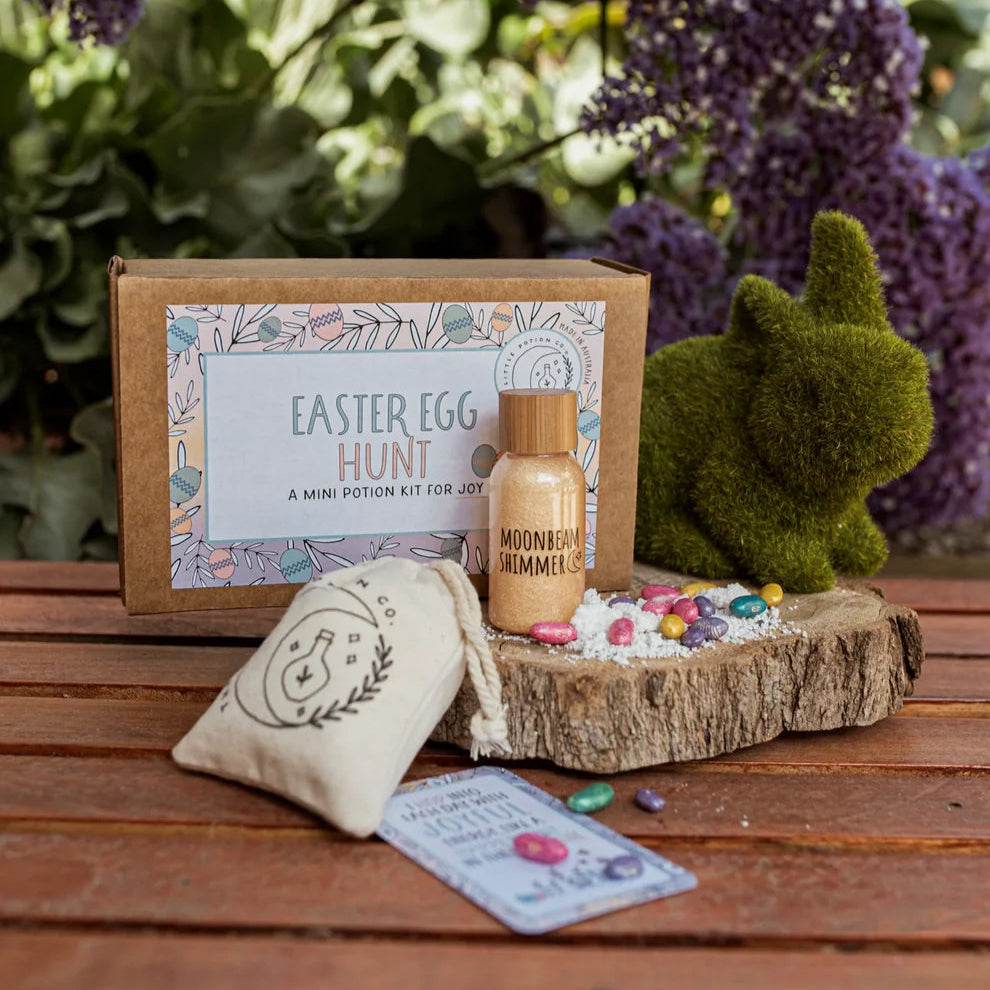 An Easter Egg Hunt Potion Kit displayed outdoors, including a cotton bag, a bottle with a liquid ingredient, a glass jar with powder, and a spell & affirmation card.