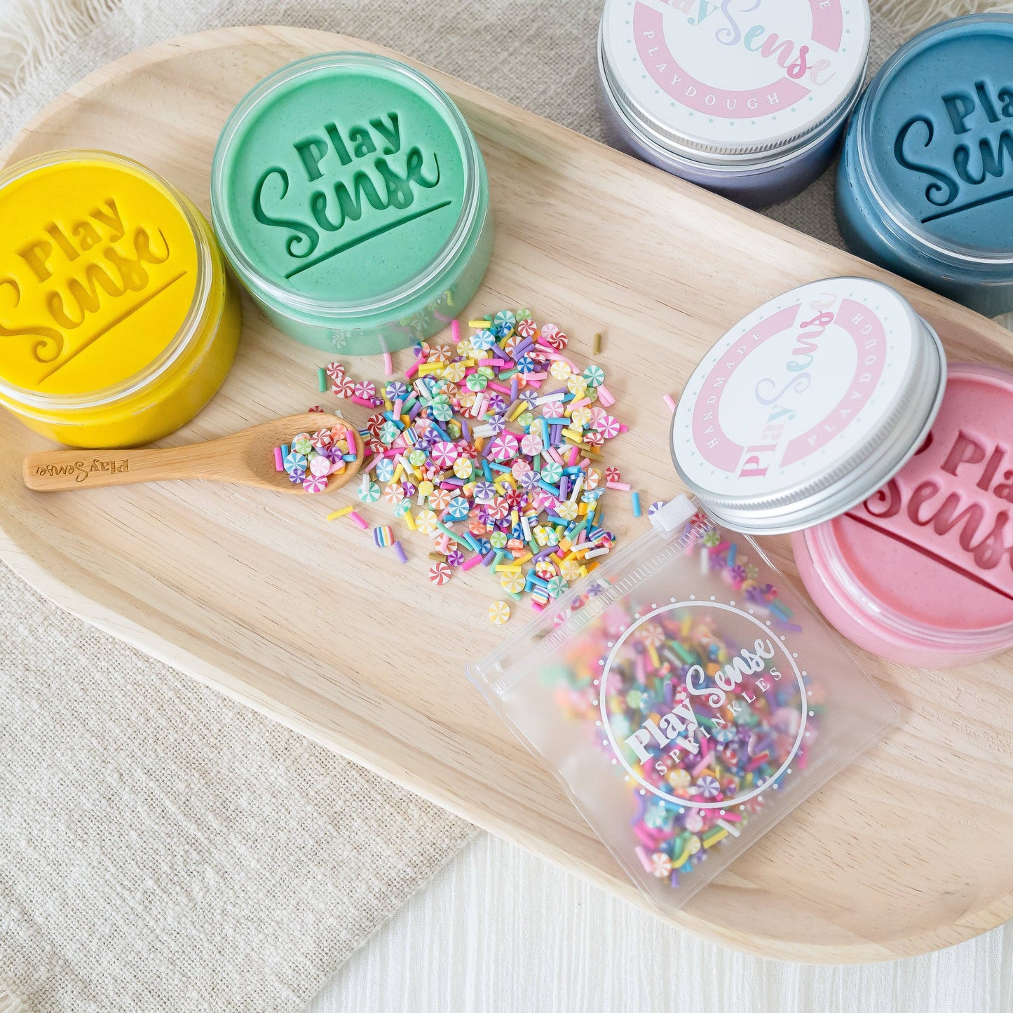 Multiple containers of Play Sense playdough with sprinkles in various colors, displayed on a wooden tray. The sprinkles are scattered in front of the containers.