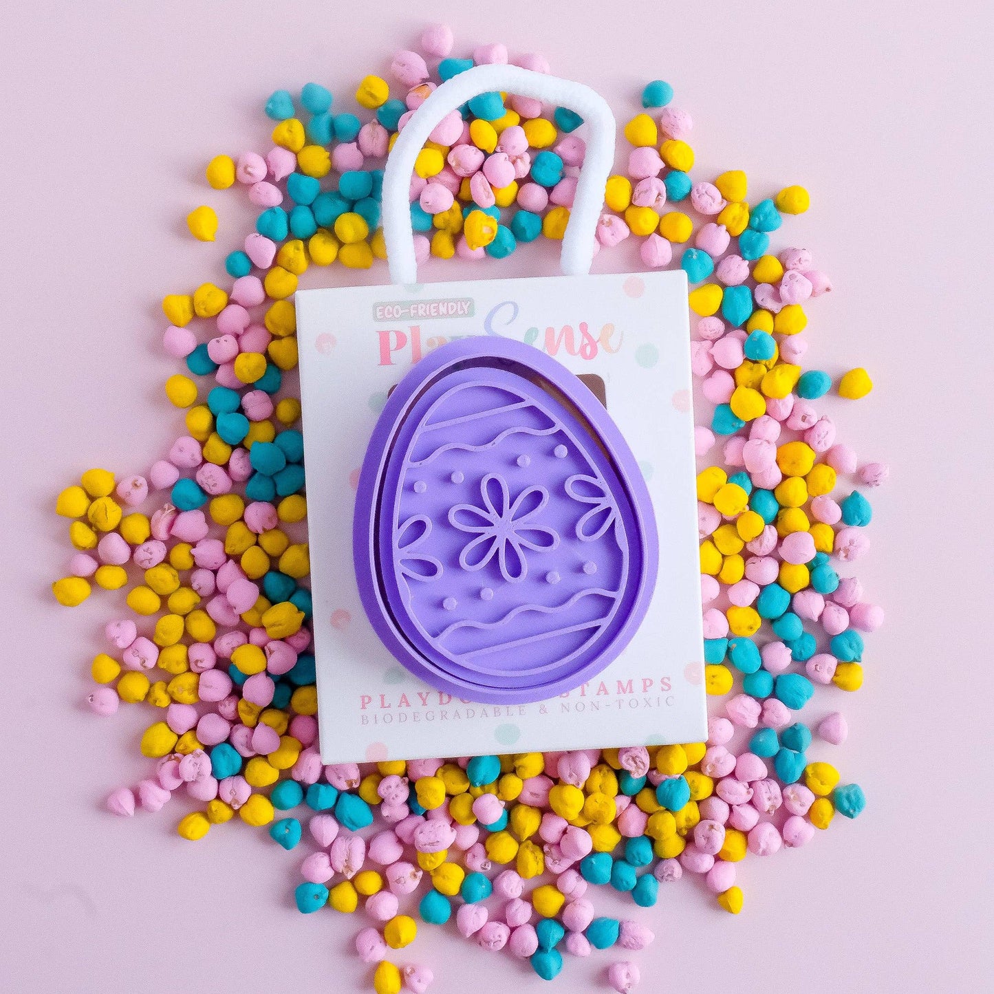 An Easter egg-shaped playdough stamp in purple color, with a handle, displayed on a surface scattered with colorful playdough.