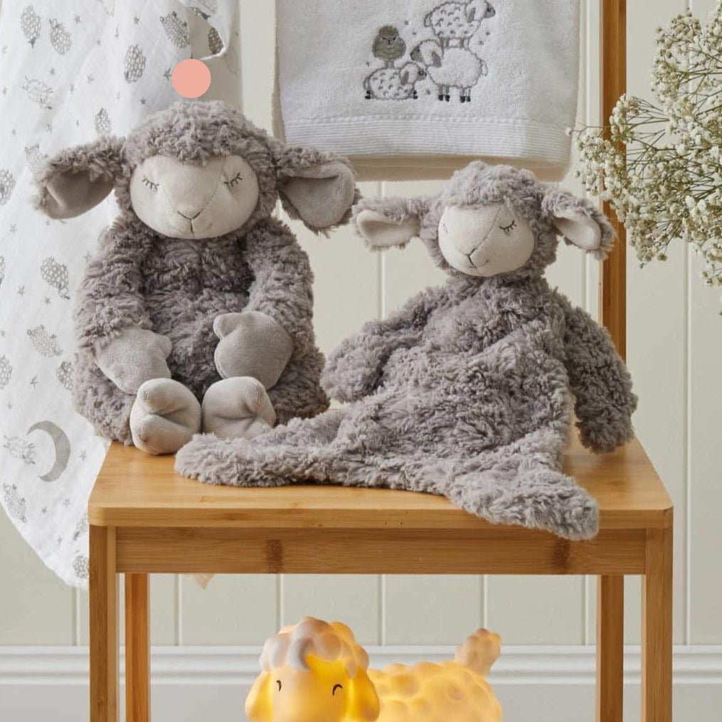 Two plush sheep toys with floppy arms and legs, sitting on a wooden table. One sheep is gray, and the other is a lighter shade, both with embroidered details and a soft, fluffy texture.