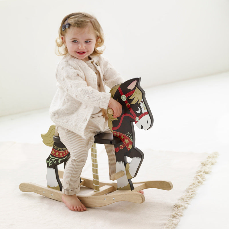 A young child riding a traditional wooden rocking horse with brown and white color scheme, featuring hand and foot grips, and decorative carousel horse and reins artwork.