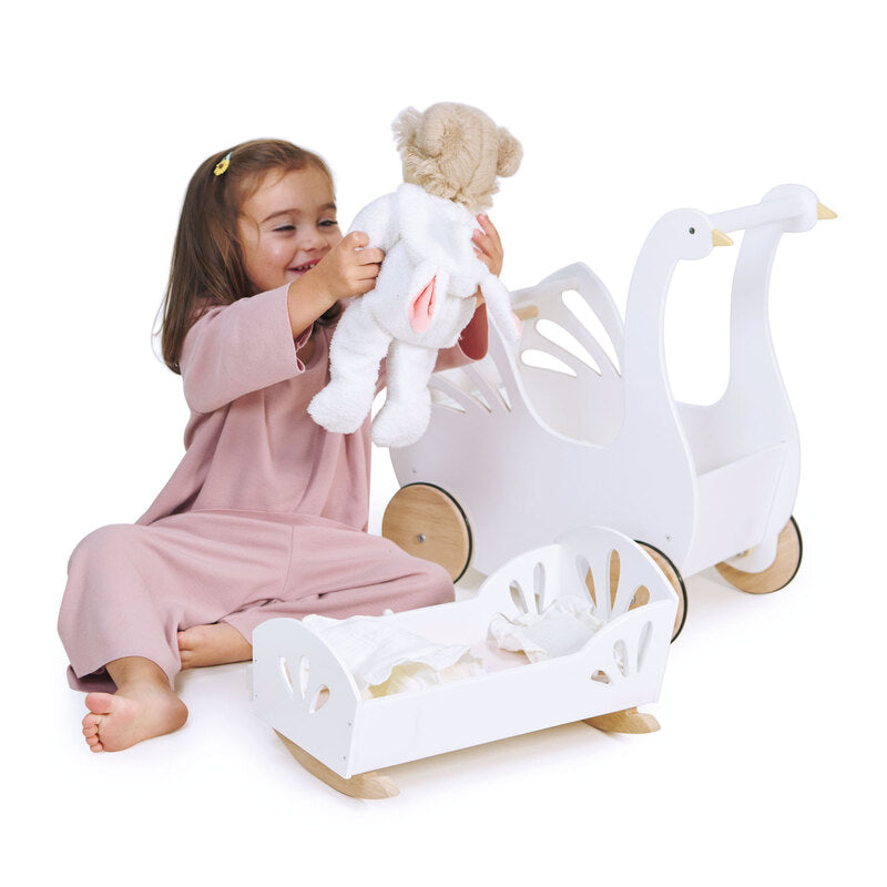 Child sitting on the floor next to a white dolls rocking cradle and swan shaped pram,  holding up a toy resembling a teddy bear.