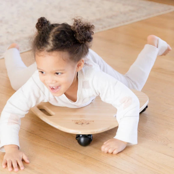 Child riding a brown wooden Tummy Glider toy with three wheels and handle cut-outs.