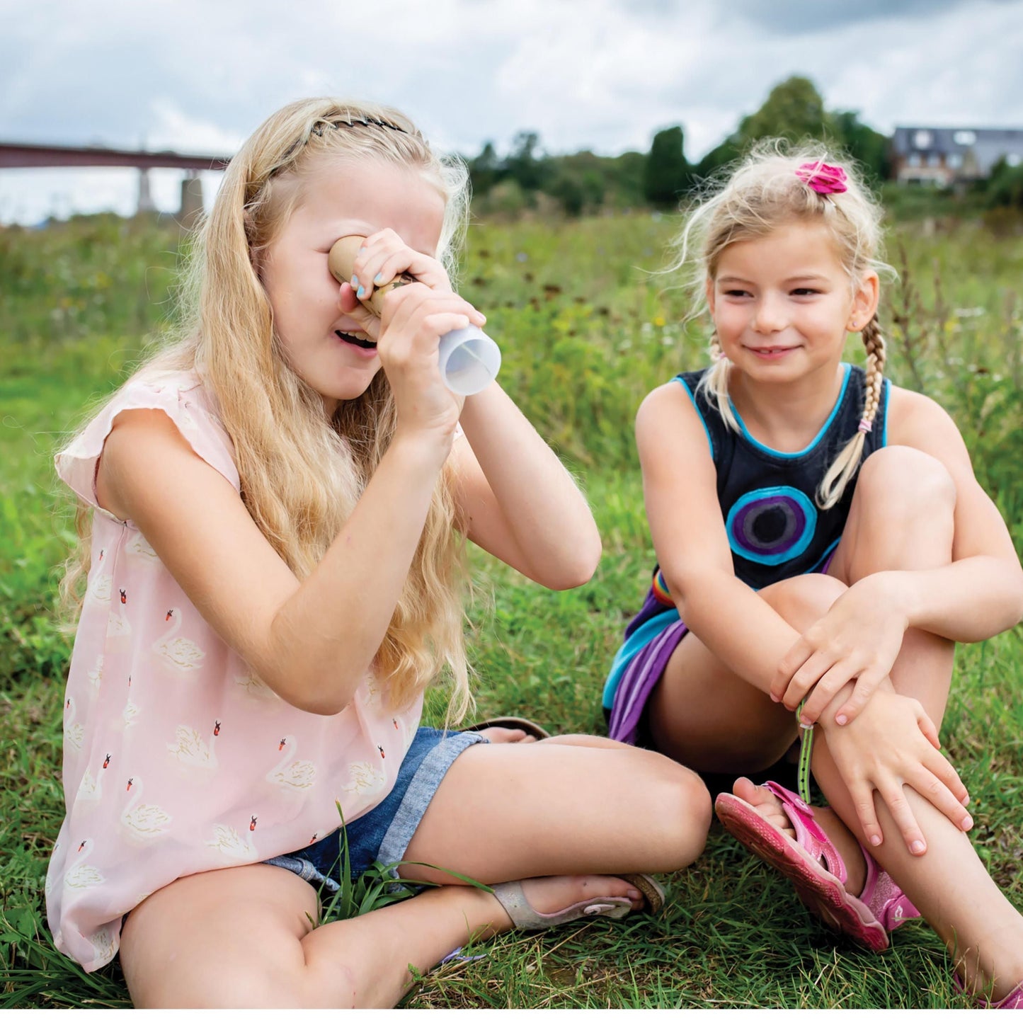 Two young girls sitting in a field, one child is looking into a kaleidoscope