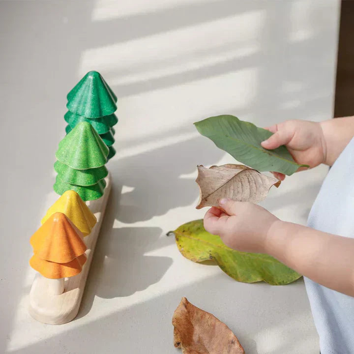 Hands of a young child holding autumn leaves next to the PlanToys tree stacking toy