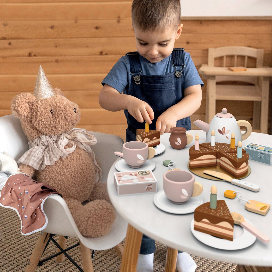 Young child having a pretend play tea party with a wooden tea set and a teddy bear wearing a party hat