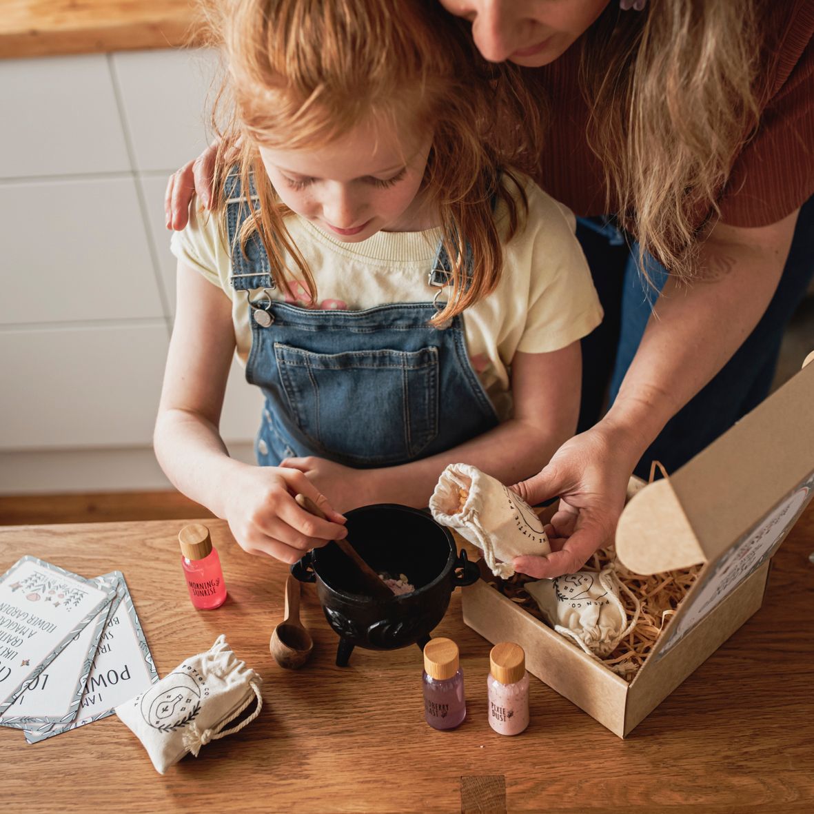 A child and an adult are exploring a box of craft materials to make potions, with various ingredients displayed on a table.