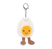 A plush toy resembling a boiled egg with a cream-colored body, yellow yolk, and brown cordy legs, attached to a keyring with a round metal Jellycat logo disc.