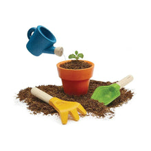 A children's gardening set including a red flower pot, a blue watering can, a green trowel, and a yellow hand fork on a pile of brown soil.