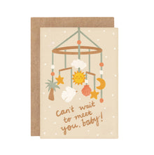 A beige greeting card with a baby hanging mobile design including stars, moon, sun, shells, a palm tree, and a bird. The text on the card says 'Can't wait to meet you, baby!' and it comes with a brown envelope.
