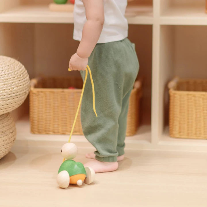 Child playing with a wooden toy duck pull-along in a room with wooden shelves and baskets.
