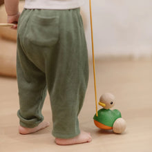 Child wearing green pants with a wooden toy on a light wooden floor.