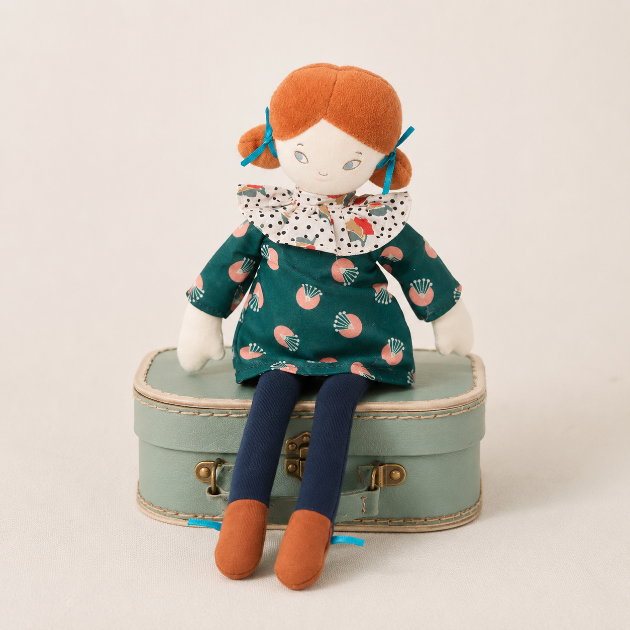 A plush doll with orange hair and a patterned green dress sits on a small mint-green suitcase against a plain light background.