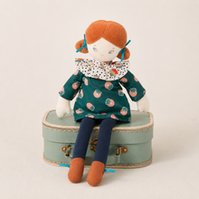 A plush doll with orange hair and a patterned green dress sits on a small mint-green suitcase against a plain light background.