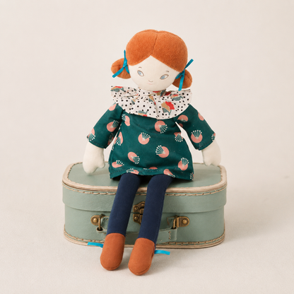 A plush doll with orange hair and a patterned green dress sits on a small mint-green suitcase against a plain light background.