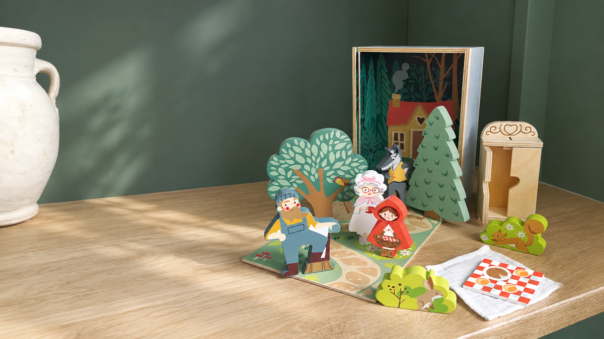 A wooden children's storytelling set displayed on a light wood table against a dark green wall. The set includes a small illustrated wooden house scene, paper-cut characters, green trees, a small wooden door, and a patterned rug or mat.