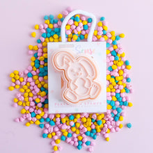 A bunny-shaped playdough stamp cutter, with a white handle and pink body, is displayed on a pink background surrounded by multicolored playdough beads.
