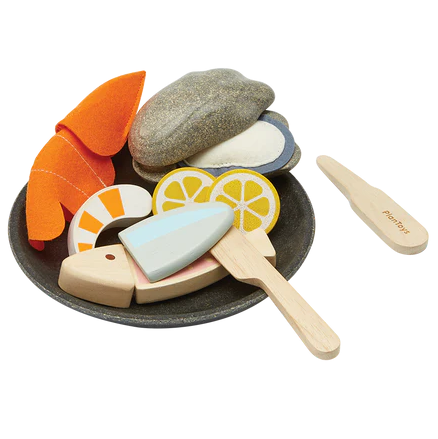 Wooden toy set resembling a seafood platter with pieces of fish, shrimp, oyster, and lemon slices, along with utensils and a plate.