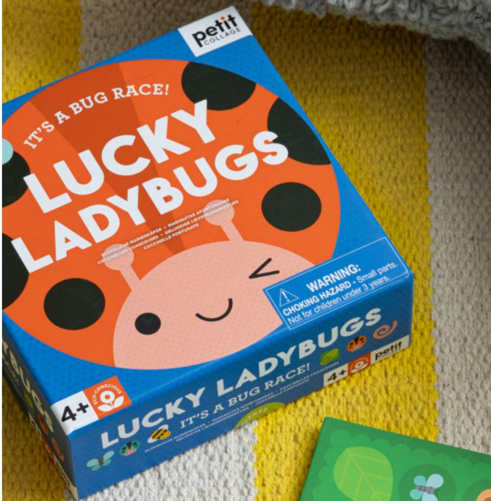 The image shows a boxed board game called 'Lucky Ladybugs' with a brightly colored ladybug on the front, along with a game die and a card, placed on a knit fabric with a yellow and white striped background.