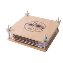 A wooden flower press made of plywood with metal screws and white sheets of paper, featuring a floral design etched into the wood.