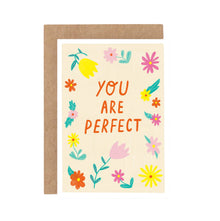 A greeting card with the text 'YOU ARE PERFECT' surrounded by colorful floral illustrations, set upon a beige background. A brown envelope is included.