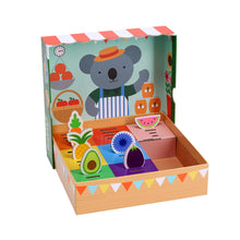 Children's educational game packaging with illustrations of colorful fruits and a koala bear. The box shows a spinner and market baskets.