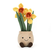 A plush toy representing a beige pot with smiling features, containing yellow and orange daffodil flowers with green leaves.