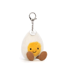 A plush toy resembling a boiled egg with a cream-colored body, yellow yolk, and brown cordy legs, attached to a keyring with a round metal Jellycat logo disc.