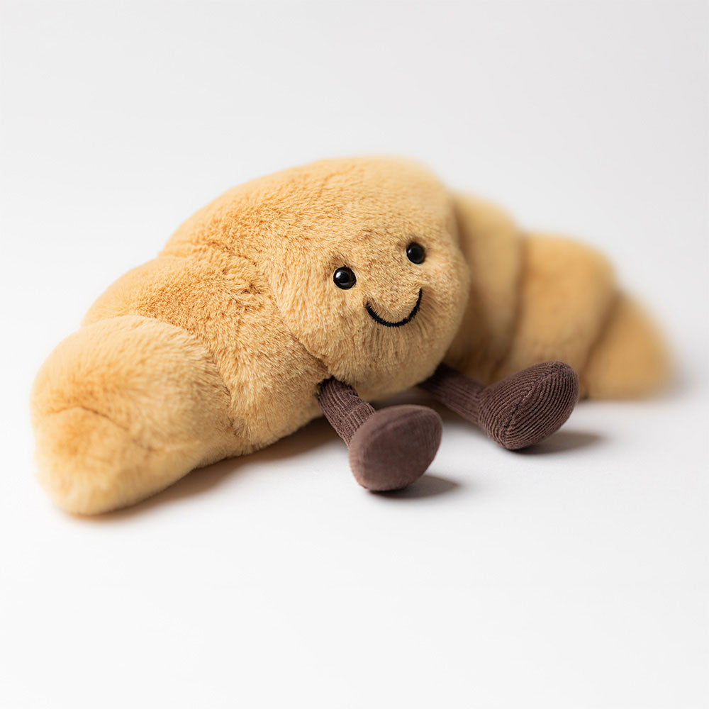 Jellycat plush toy resembling a croissant with a smiling face on a white background