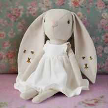 Stuffed toy in the shape of a bunny with a white floral dress and embroidered details, including bees on the skirt. Sitting against a pink and green floral fabric background.