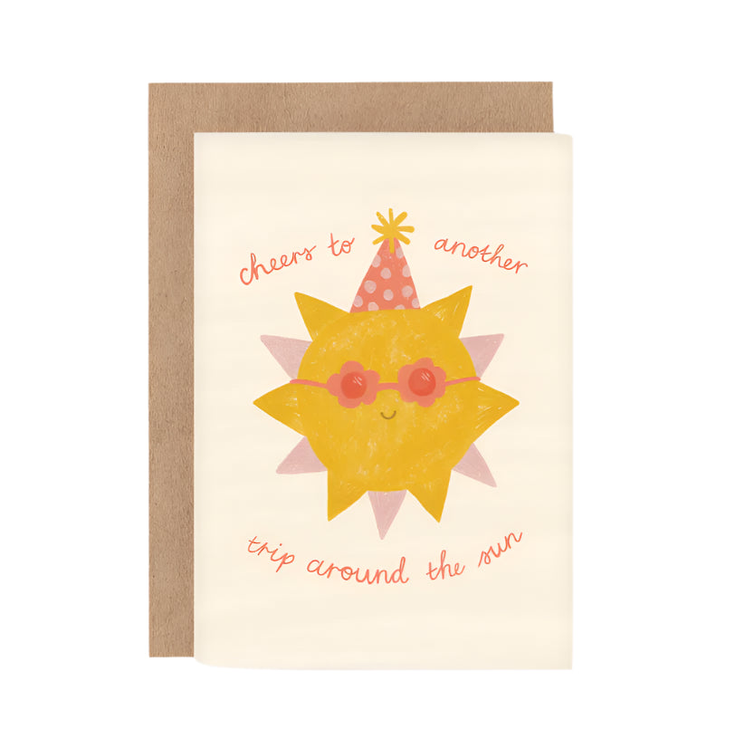 A greeting card with an illustration of a sun wearing sunglasses and a party hat, with the text 'cheers to another trip around the sun'. There is a beige background and a brown envelope included.
