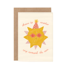 A greeting card with an illustration of a sun wearing sunglasses and a party hat, with the text 'cheers to another trip around the sun'. There is a beige background and a brown envelope included.
