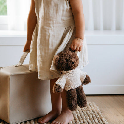 Child in a light dress holding a teddy bear next to a suitcase indoors.