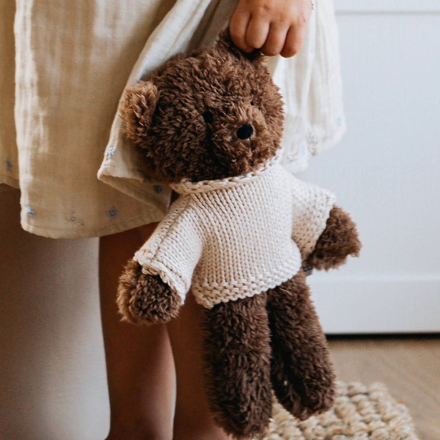 Brown teddy bear wearing a white sweater held by a person in a neutral setting