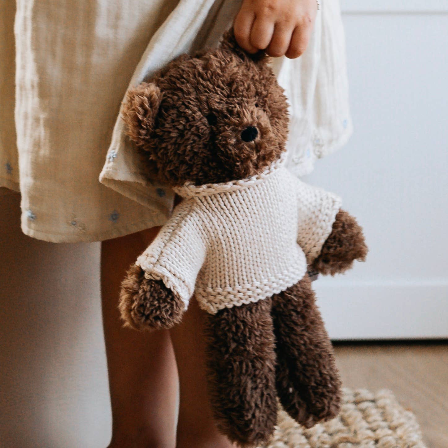 Brown teddy bear wearing a white sweater held by a person in a neutral setting