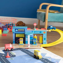 A wooden toy service station with a garage, ramp, pumps, and parking accessories, along with a few toy cars and a bear figure in the background.