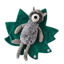A plush toy of a bedtime baby raccoon with a soft gray body and a green leaf-shaped blanket.